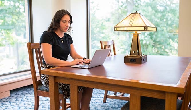 A student uses their laptop at a desk surrounded by windows.