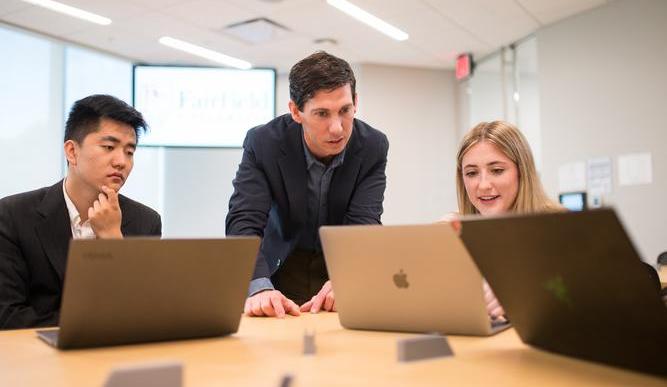 Two students work at laptops at a table, one is explaining something to a professor who looks at her screen