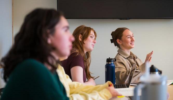 Three students sit in a classroom, one is talking while the others listen. 