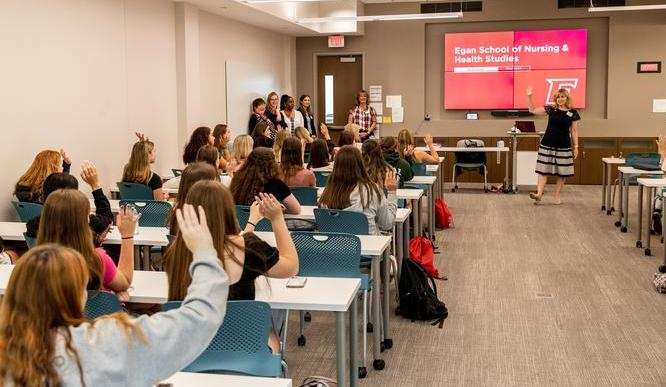 A classroom of students sit at desks raising their hands in front of professors and a screen that reads “Egan School of Nursing and Health Studies”.