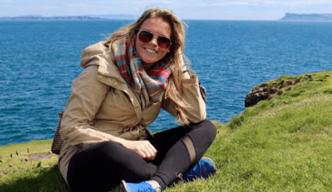 A student sits on a grassy cliff overlooking bright blue water with a horse in the background.