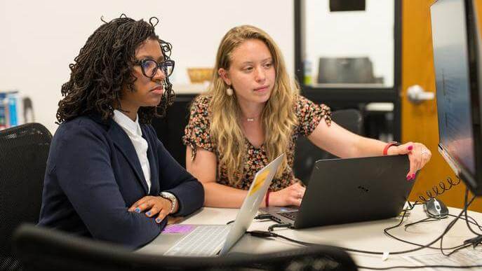 Two graduate students work together in an office with laptops. One points at a computer screen while the other looks focused. 