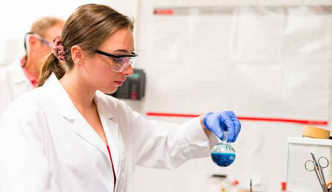 A woman in a lab coat holds a beaker filled with a vibrant blue liquid, showcasing her work in a laboratory setting.