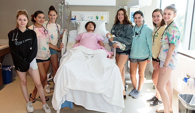 Pre-college, prospective Nursing students smiling in a simulation environment with a sim-doll patient.