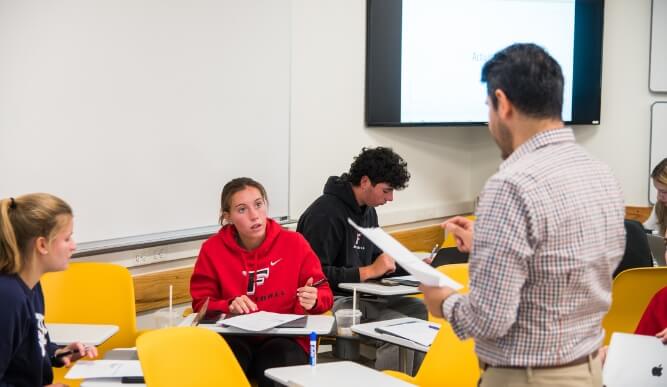 A professor holding a piece of paper addresses students in a classroom.