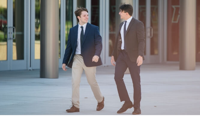 Two students wearing suits walking and talking in front of a building on campus.