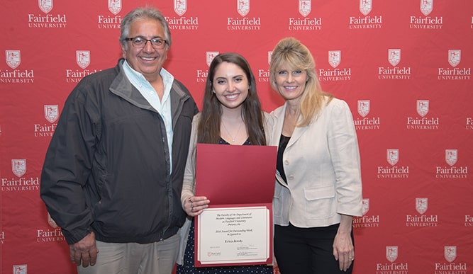 A student surrounded by her parents holds a certificate in front of a red Fairfield background.